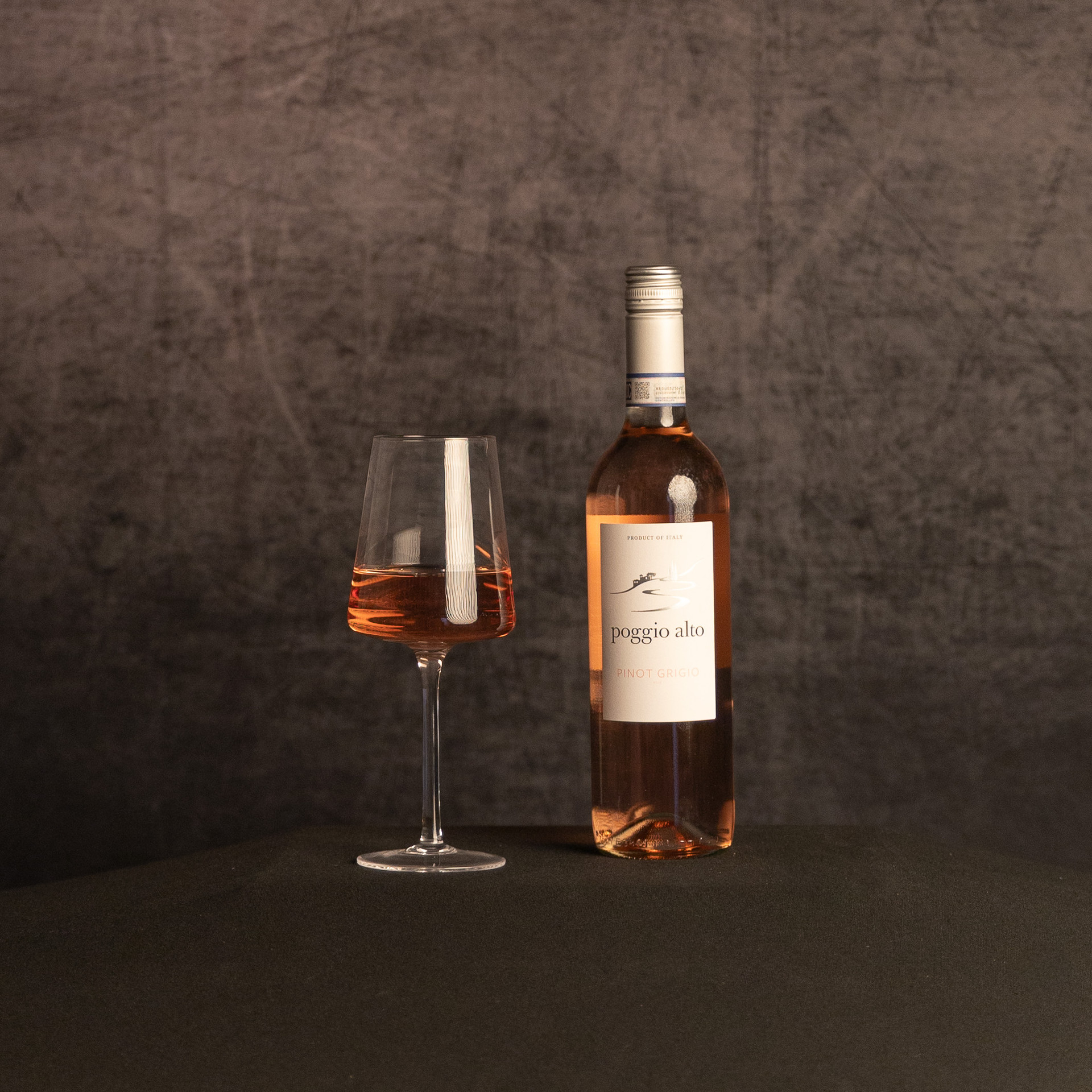 A bottle of Poggio Alto Pinot Grigio Rosé beside a partially filled glass of rosé wine, photographed against a dark, textured brown backdrop.