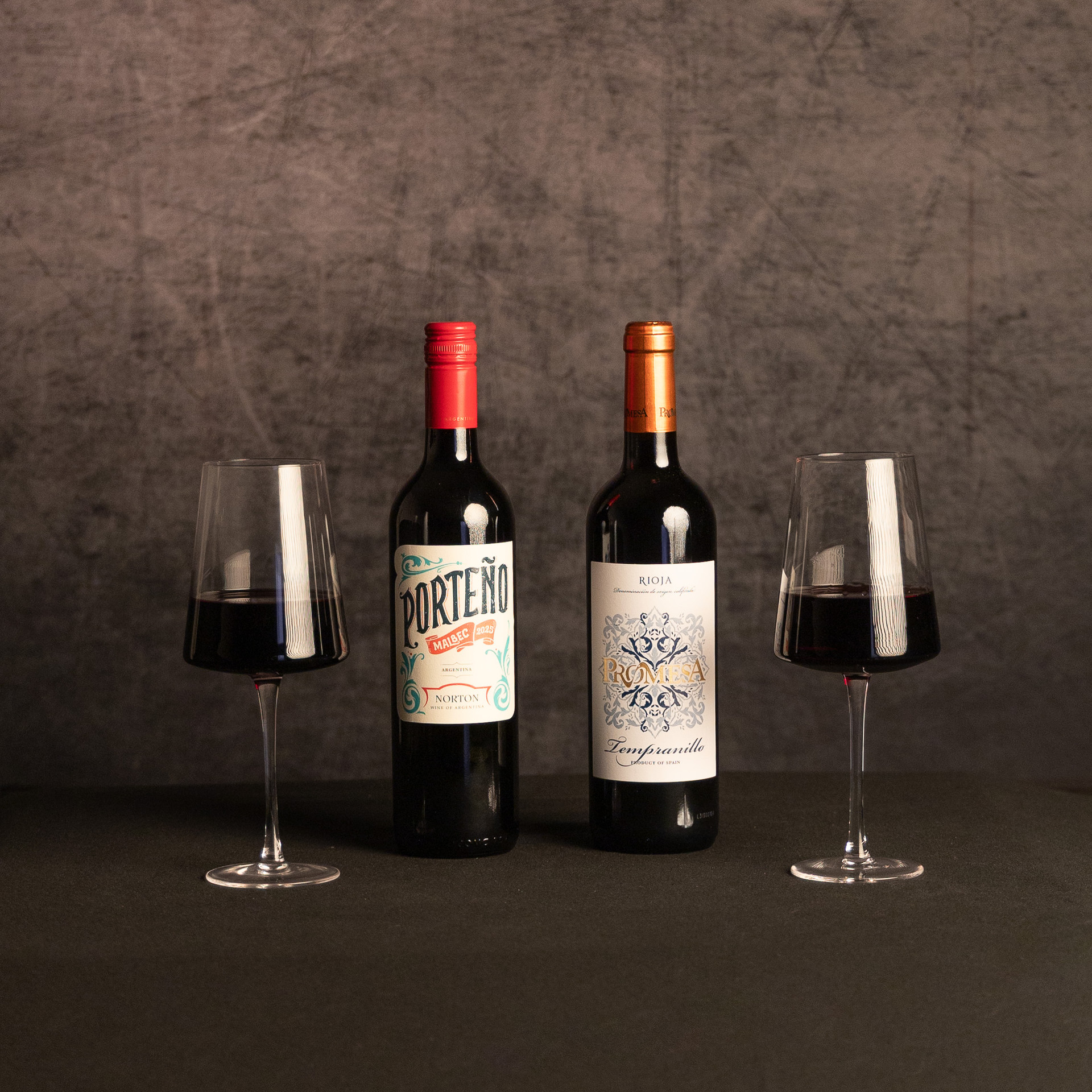 Bottles of Porteño Malbec and Promesa Rioja Tempranillo stand between two glasses of red wine against a dark, textured backdrop.
