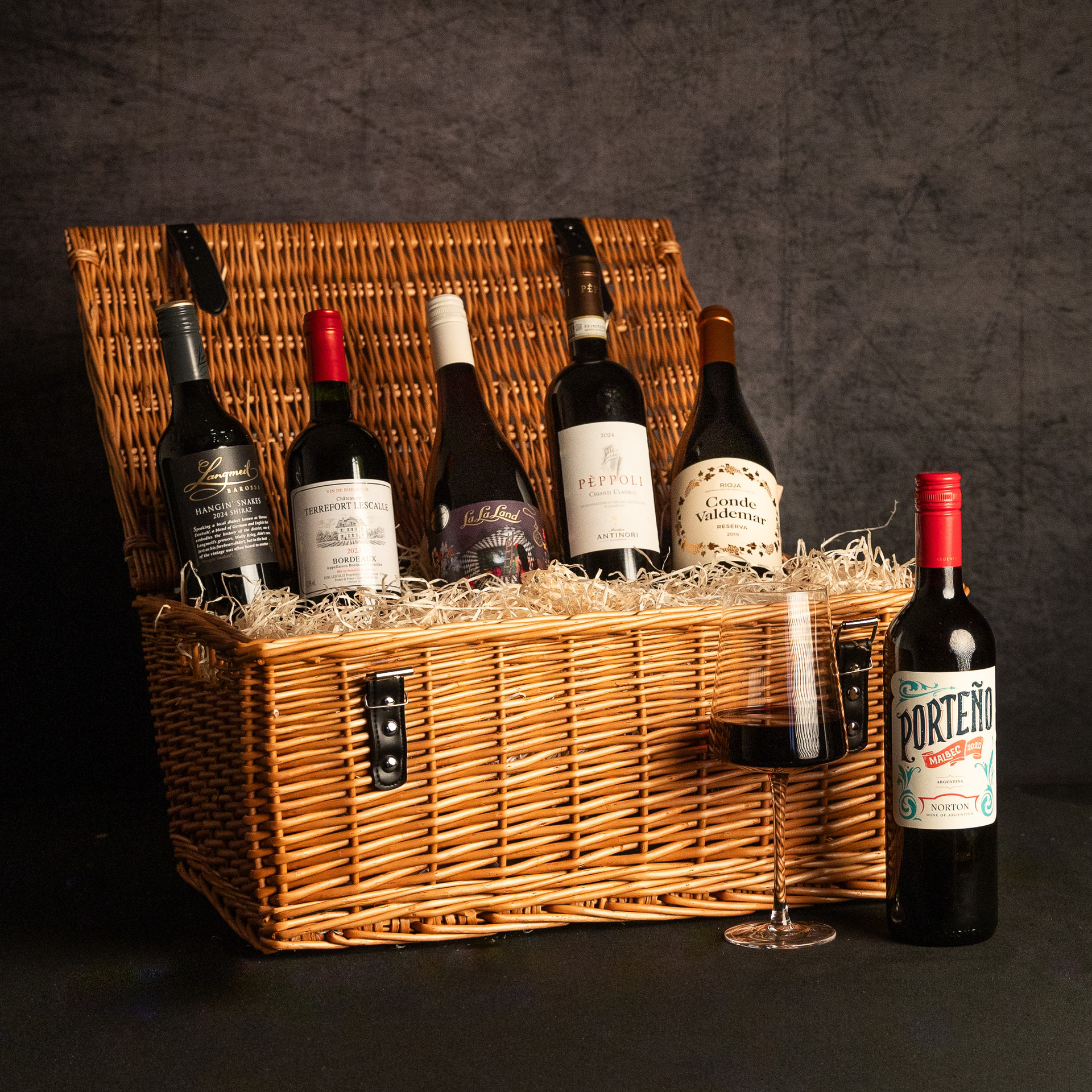 An open wicker hamper filled with five bottles of red wine, including Langmeil Hanging Snakes Shiraz, Château Terrefort Lescalle Bordeaux, La La Land Pinot Noir, Pèppoli Chianti Classico and Conde Valdemar Reserva, with a bottle of Porteño Malbec and a glass of red wine alongside.