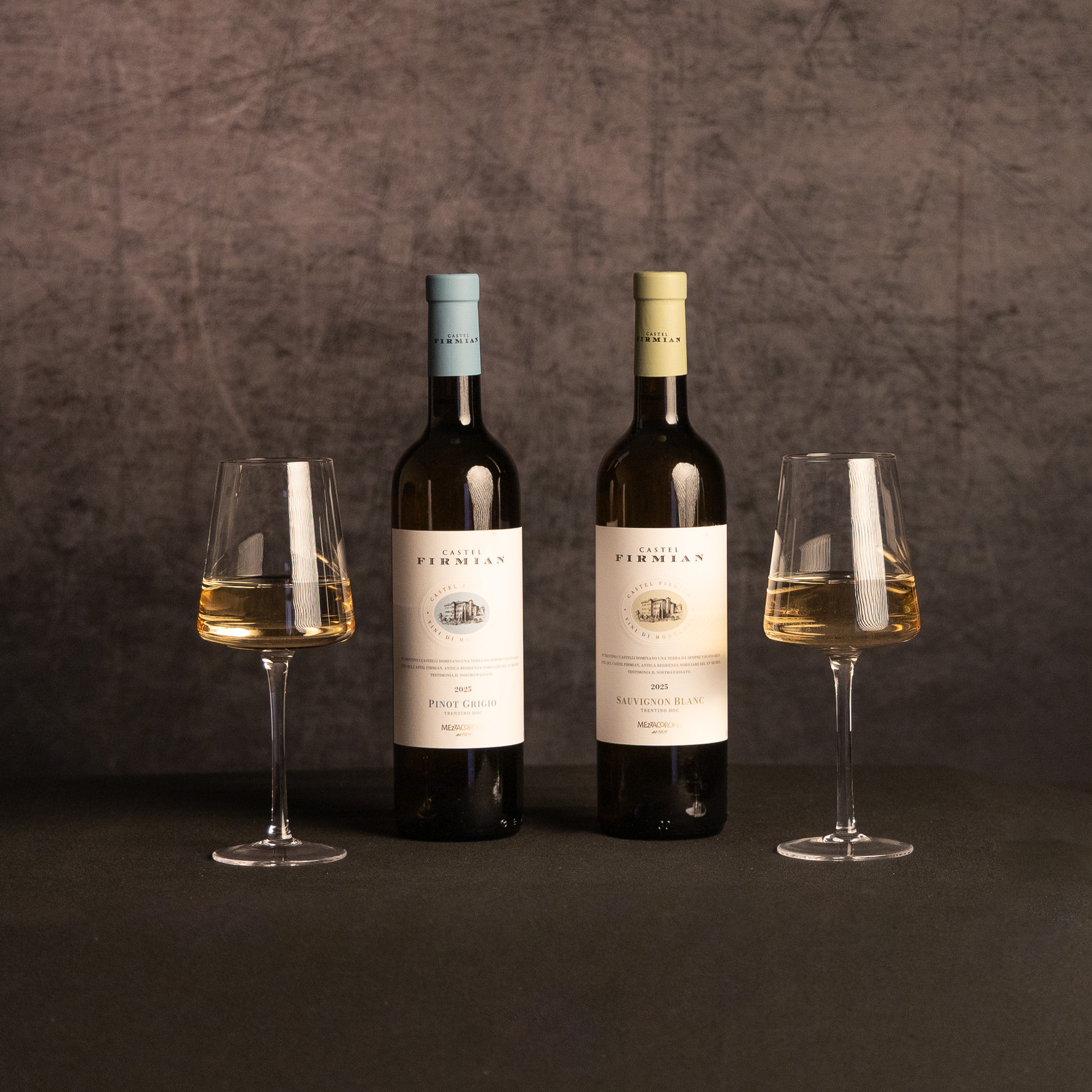 Two bottles of Castel Firmian white wine—Pinot Grigio and Sauvignon Blanc—stand between glasses of pale white wine against a dark, textured backdrop.