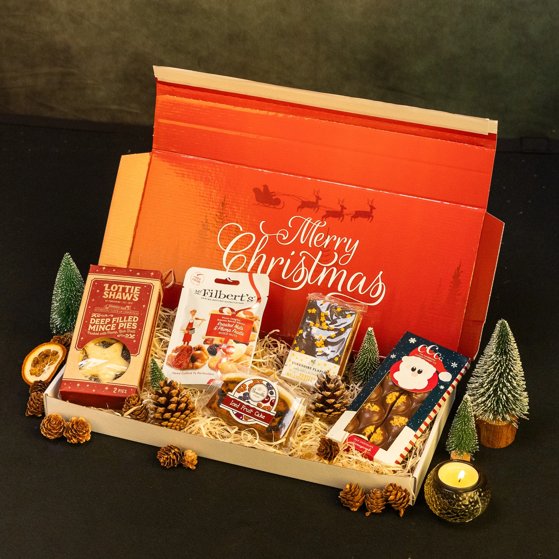 Open Christmas hamper box filled with festive treats, including mince pies, mixed nuts, fruit cake, chocolate flapjack and chocolate sweets, surrounded by miniature Christmas trees, pinecones, dried orange slices and a lit candle.