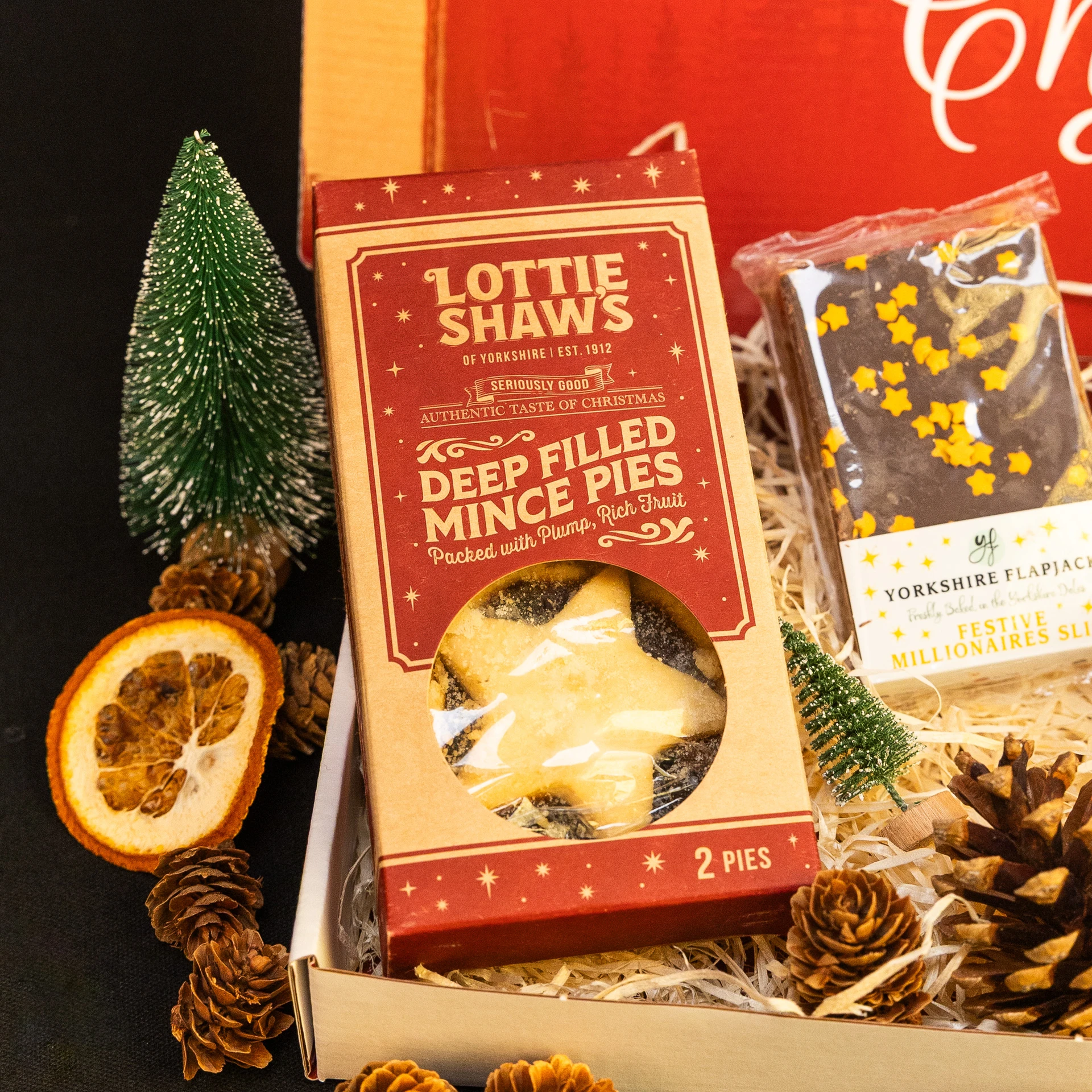 Close-up of Lottie Shaw’s deep-filled mince pies in festive red packaging, displayed inside a Christmas hamper alongside a chocolate flapjack, pinecones, a miniature Christmas tree and a dried orange slice.