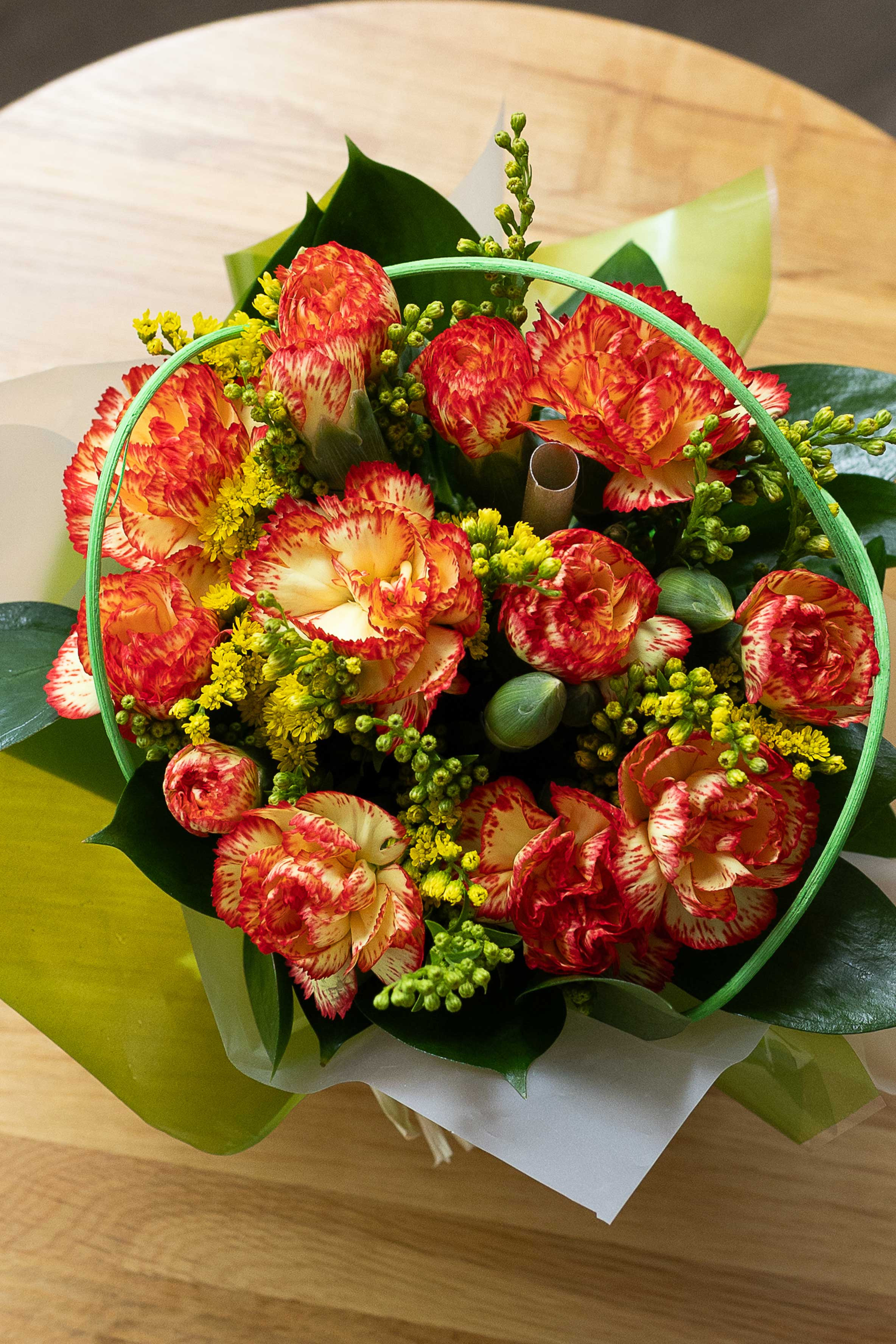 Top-down view of a posy of orange-and-yellow carnations with yellow filler flowers and green foliage on a wooden table.