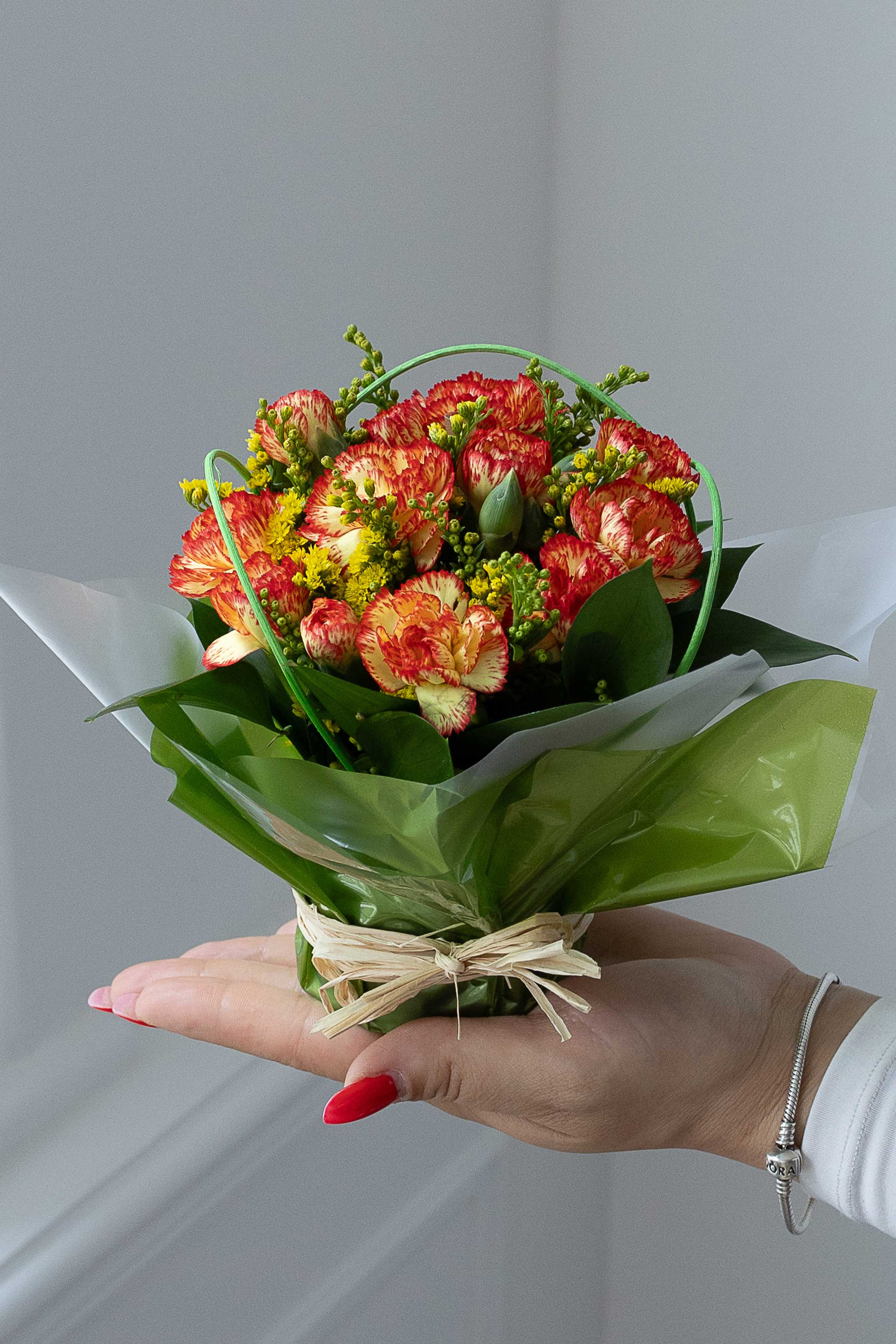 Hand holding a posy of orange and yellow carnations with yellow filler flowers, wrapped in green and white paper and tied with raffia.