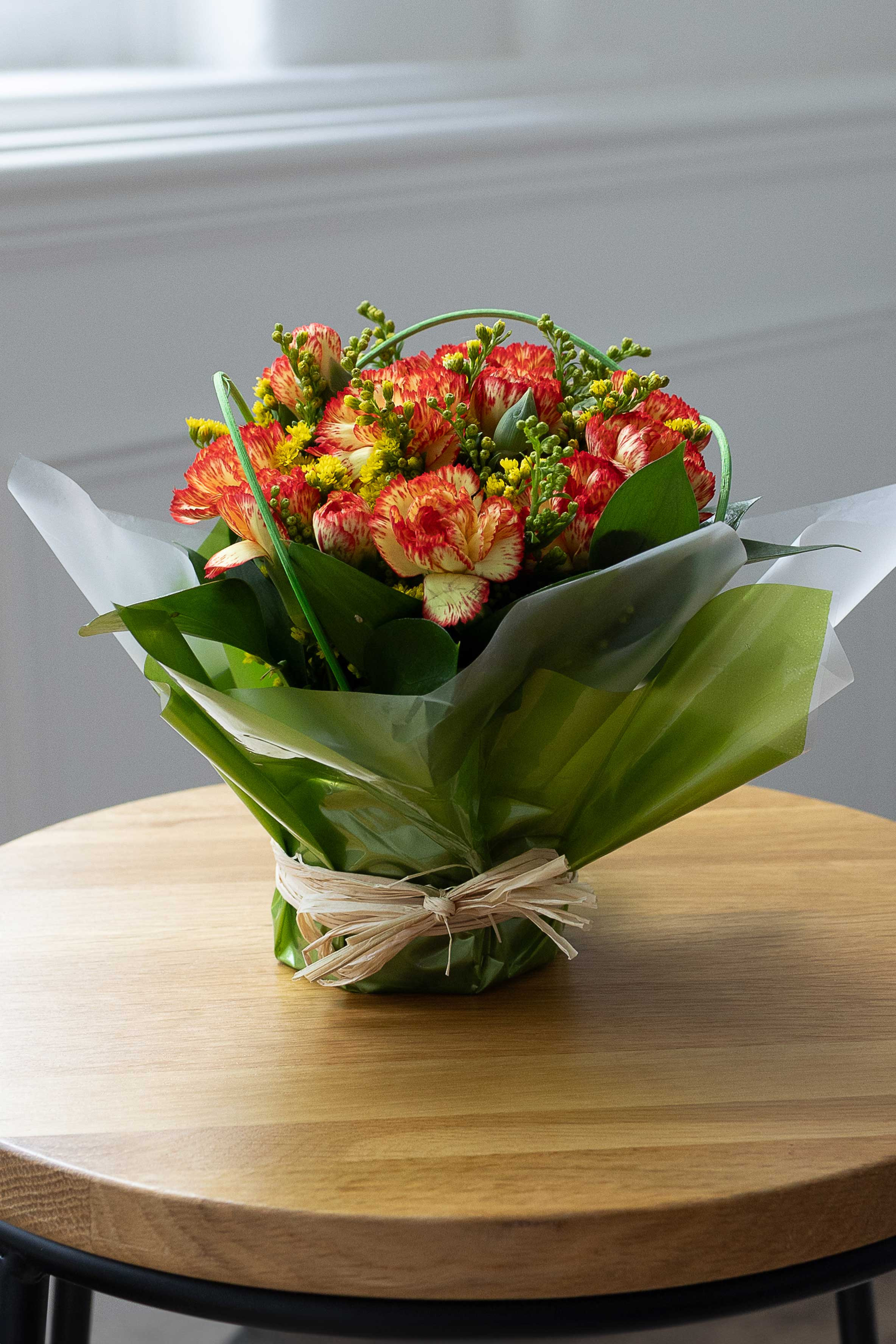 Orange-and-yellow carnation posy with yellow filler flowers, wrapped in green and white paper and displayed on a wooden table.