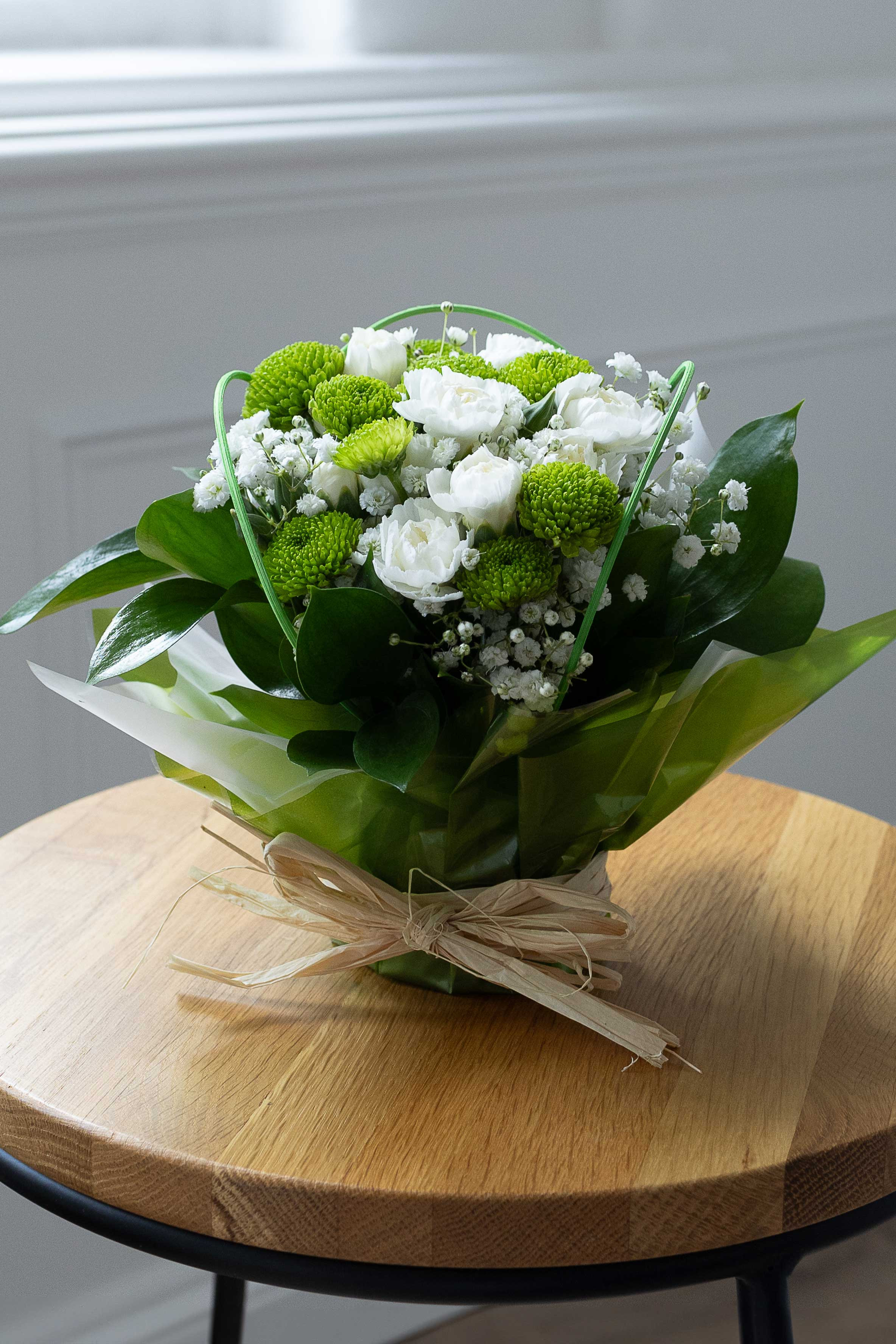 Green button chrysanthemums, white lisianthus, and baby's breath posy wrapped in green and white paper on a wooden table.