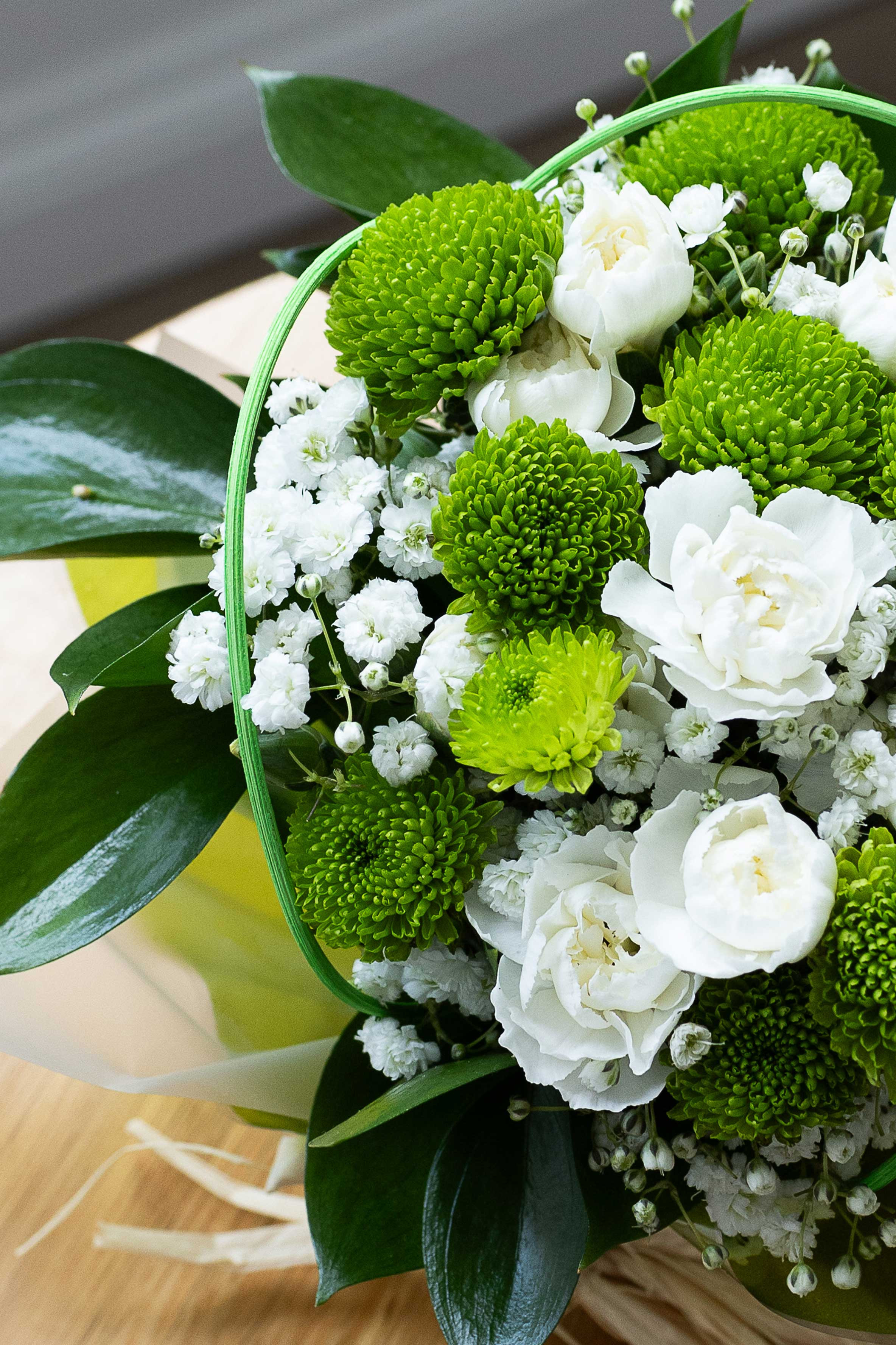 Close-up of green button chrysanthemums, white lisianthus, and baby's breath arranged in a posy with dark green foliage.