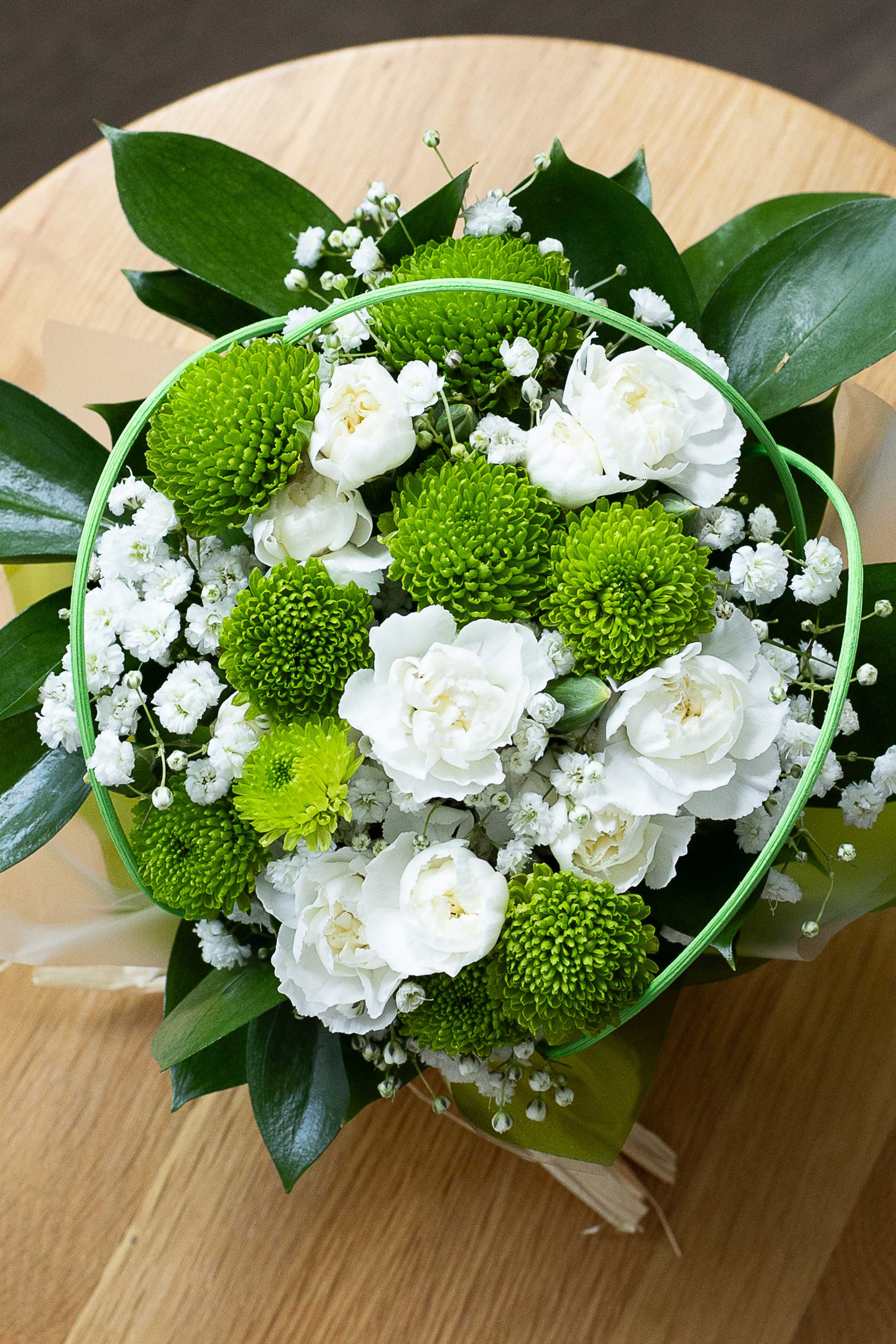 Top-down view of a posy of green button chrysanthemums, white lisianthus, and baby's breath with green foliage on a wooden table.