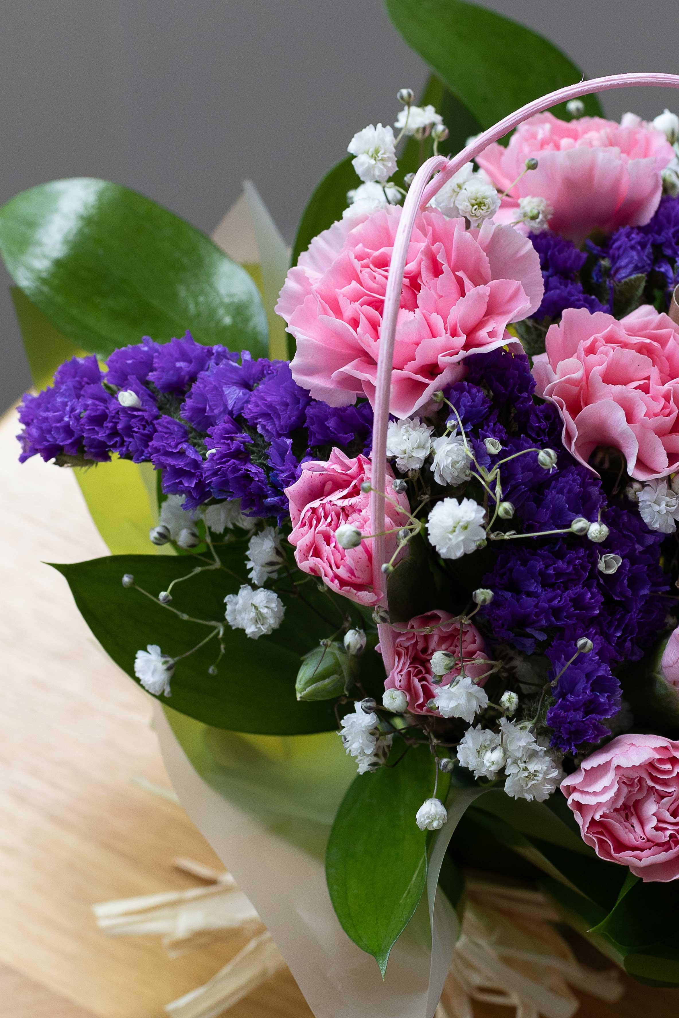 Close-up of pink carnations, purple statice, and white baby's breath arranged in a posy with green foliage.
