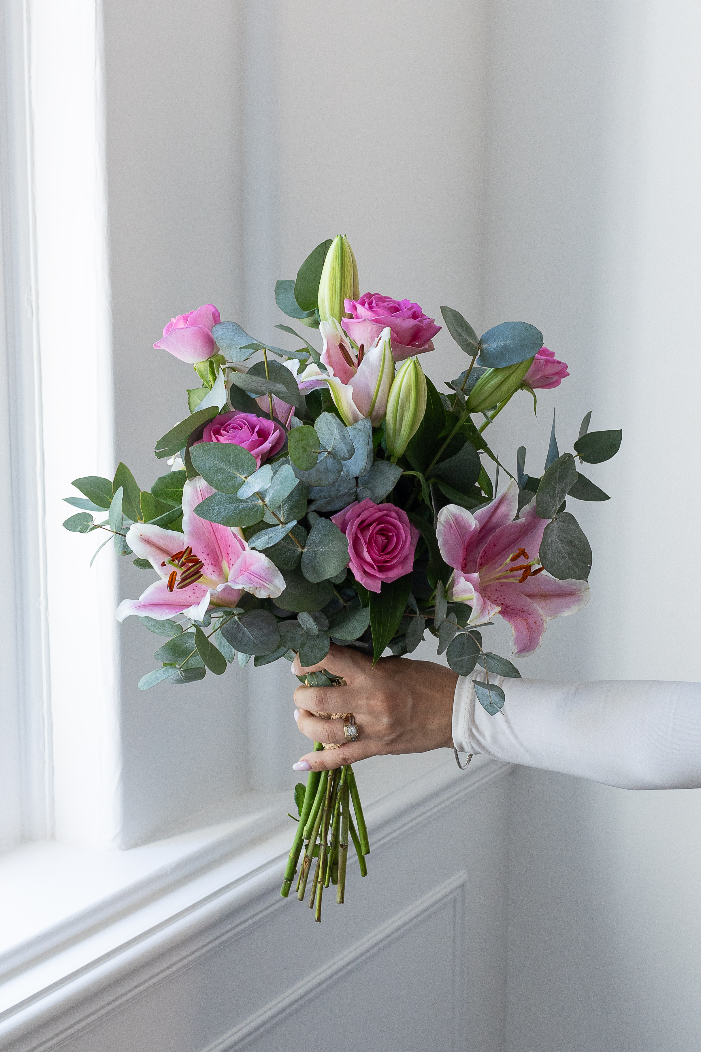 Hand holding a bouquet of pink roses, pink lilies, and eucalyptus beside a bright window.