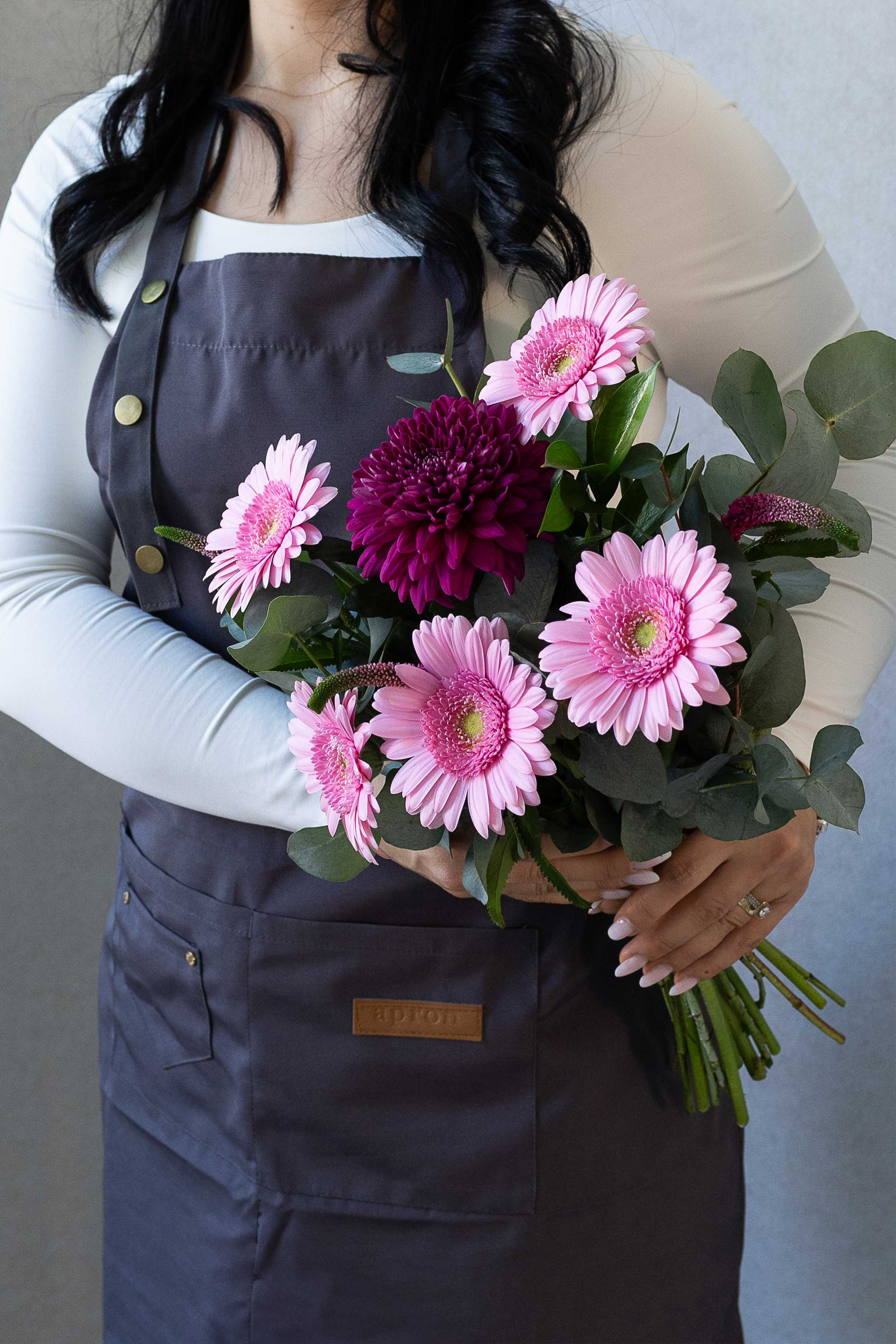 Florist holding a bouquet of pink gerbera daisies, a burgundy chrysanthemum, eucalyptus, and veronica flowers.