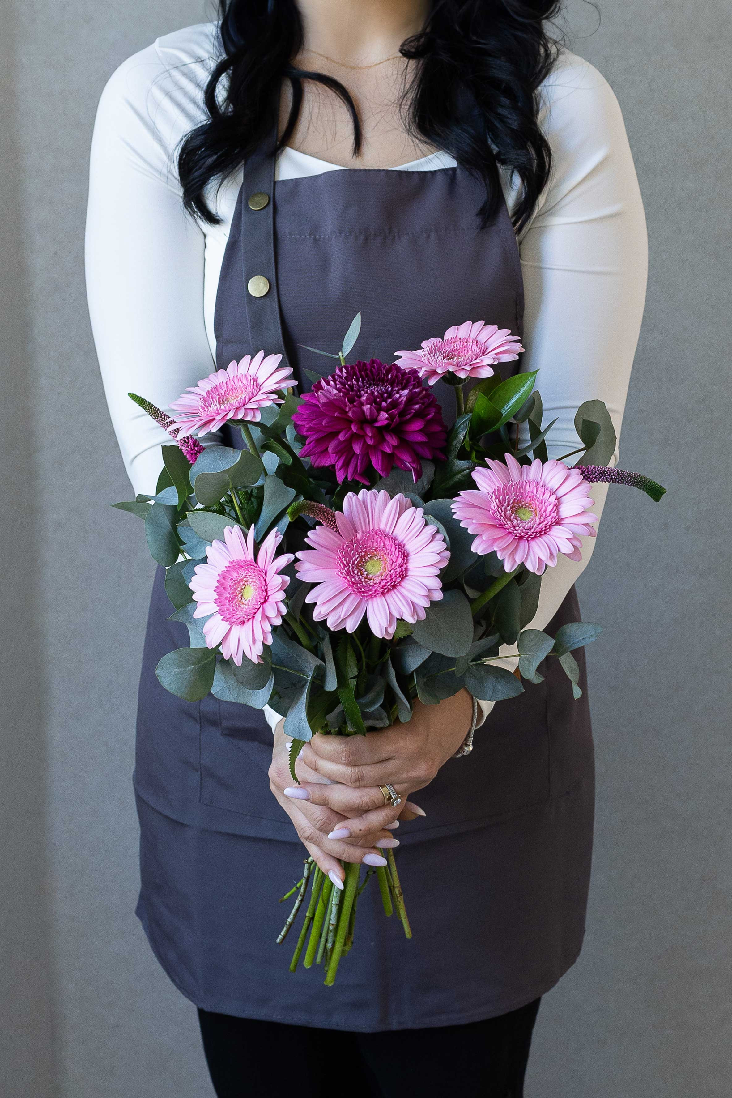 Florist holding a bouquet of pink gerbera daisies, a burgundy chrysanthemum, eucalyptus, and veronica flowers against a gray background.