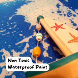 Non-toxic waterproof painted wooden sailboat with colourful beads on the mast string, set against a blue sea-inspired background.