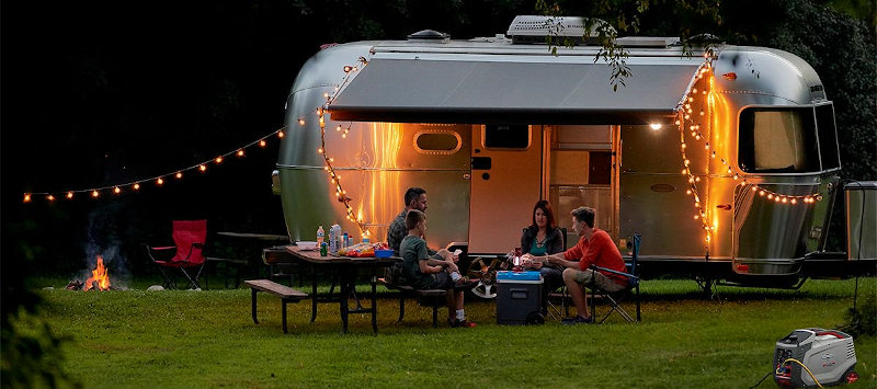 A Briggs &* Stratton portable inverter generator powers family fun at an RV campsite. The family has light strings and outside by the table. Inside, a rooftop air conditioner keep the trailer RV cool and comfortable.