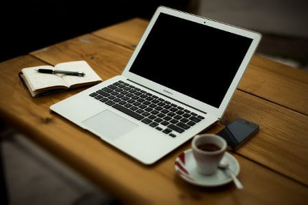 A Macbook Laptop on a desk with coffee,  notebook, and smart phone.