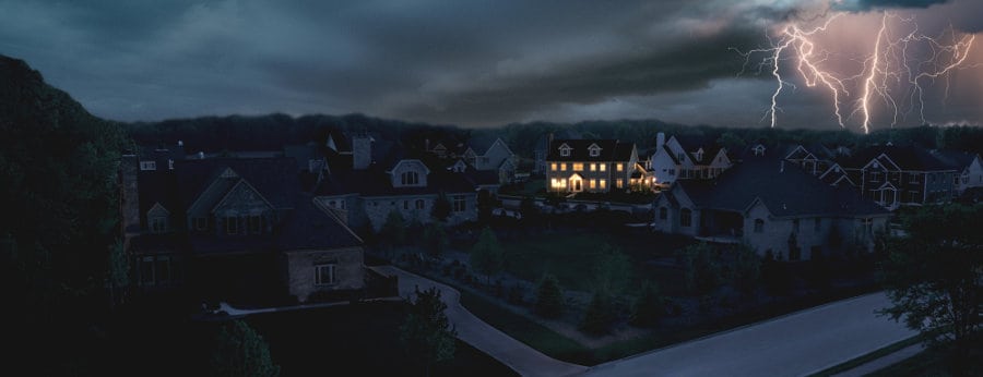 A single home has lights in a dark neighborhood after a severe storm caused a power outage. Lightning flashes in the background highlight dark, ominous clouds