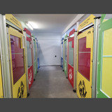 Two rows of face to face kennels with glass doors in red, lime green, royal blue and a custom yellow. The yellow kennels have lids to help contain even the craziest jumpers. Each door has their custom logo and custom headers which displays that kennel sponsor.  The bright colored kennels really pop compared to the grey concrete floor and white walls adding a visual appeal.