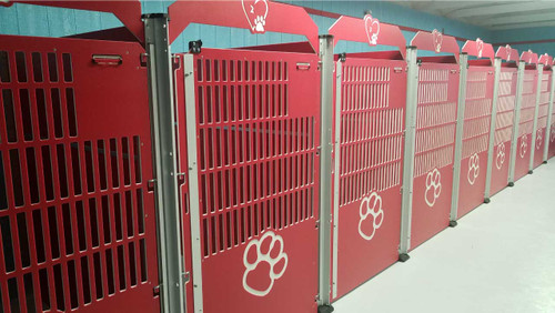 Row of red dog kennels. Each kennel gate has a pawprint design on it. The bottom portion of the gates are solid to reduce fence-fighting, while the upper portion of the gates are slotted for ventilation and visualization into the kennels. A heart design is engraved above each gate with a number inside of it. A built-in clipboard clip keeps the occupants' information right on the kennel for the staff to keep track of.