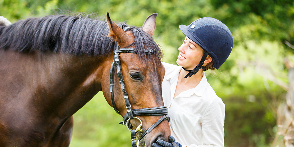 Young smiling rider woman in helmet holding bay horse by bridle