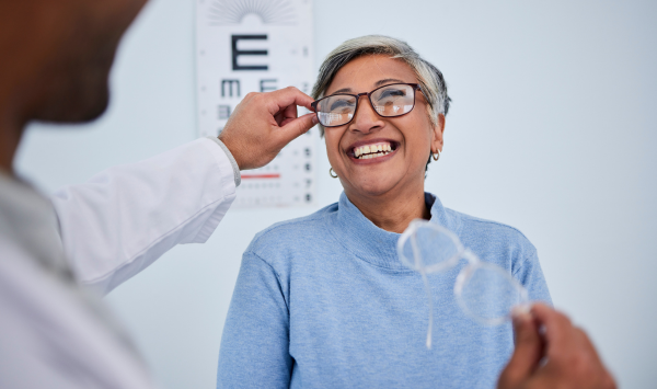 Older adults at an eye clinic, smiling and trying on glasses after an exam, representing clear vision and eye health support.