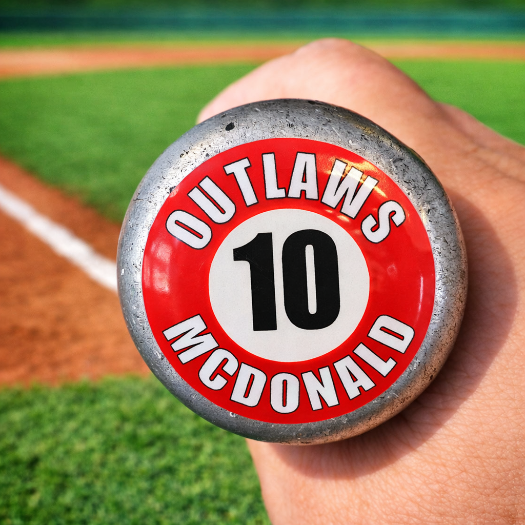 Close-up of a baseball bat knob featuring a custom personalized sticker with the player name “Outlaws McDonald” and number 10. The durable, waterproof decal is applied to the end of the bat for easy identification. The background shows a blurred baseball field with green grass and a dirt infield, highlighting the game-ready look and real-use setting.