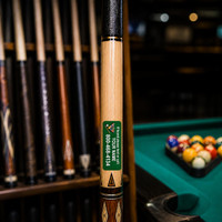 Stinky Lockers Personalized Pool Cue ID Decals, close-up of a billiard cue with custom sticker on the shaft in a pool hall with pool balls and cues in the background.