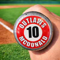 Close-up of a baseball bat knob featuring a custom personalized sticker with the player name “Outlaws McDonald” and number 10. The durable, waterproof decal is applied to the end of the bat for easy identification. The background shows a blurred baseball field with green grass and a dirt infield, highlighting the game-ready look and real-use setting.