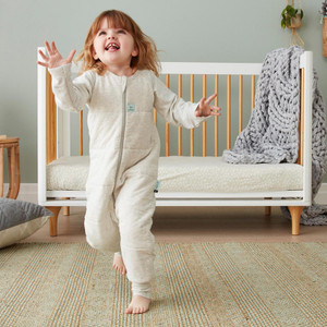 Child wearing ergoPouch grey marle organic cotton padded sleep onesie standing in modern nursery with white cot