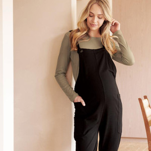Pregnant woman wearing Ripe Maternity black overalls with olive long-sleeve top, touching hair in neutral-toned room