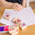Melissa & Doug Friendship Stamp Set - child using rose design wooden stamp on white paper with storage box, stamps and pencils at a wooden table | Peekaboobaby