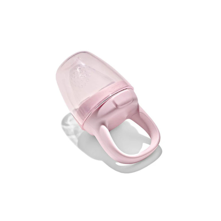 OXO Tot pink silicone self-feeder with handle and mesh pouch laying on white background