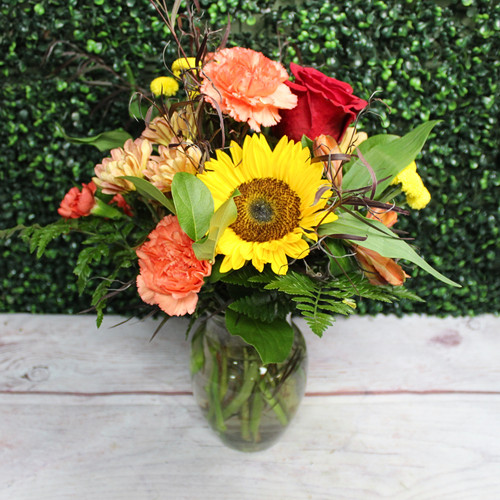 Sunflower, red rose, orange carnations, and peach mums with fern in a clear glass vase on a wood table.
