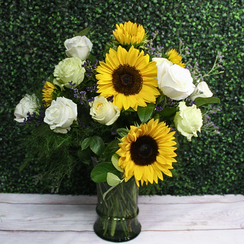 Large arrangement of yellow sunflowers and white roses with purple statice in a clear glass vase.