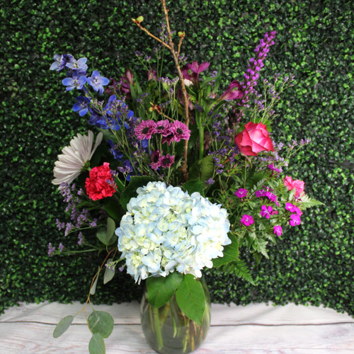 Blue hydrangea, purple roses, hot pink carnations, and blue delphinium in a clear vase