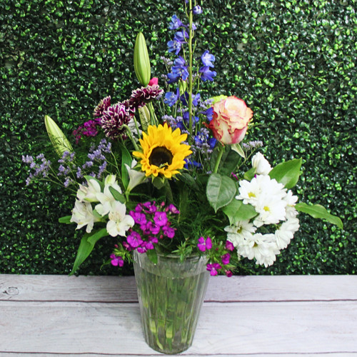 Sunflowers, stargazer lilies, delphinium, roses, and daisies in a tall clear glass vase