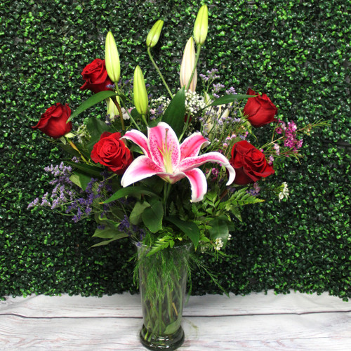 Six red roses and a pink stargazer lily with purple statice, baby’s breath, and ferns in a clear glass vase