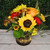 Sunflower, orange rose, red carnations, and fall mums with wheat in a dark hurricane vase with warm lights on a wood tabletop.
