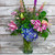 Tall arrangement of blue hydrangea, pink stargazer lilies, peach roses, and dianthus with purple liatris in a green-tinted glass vase.