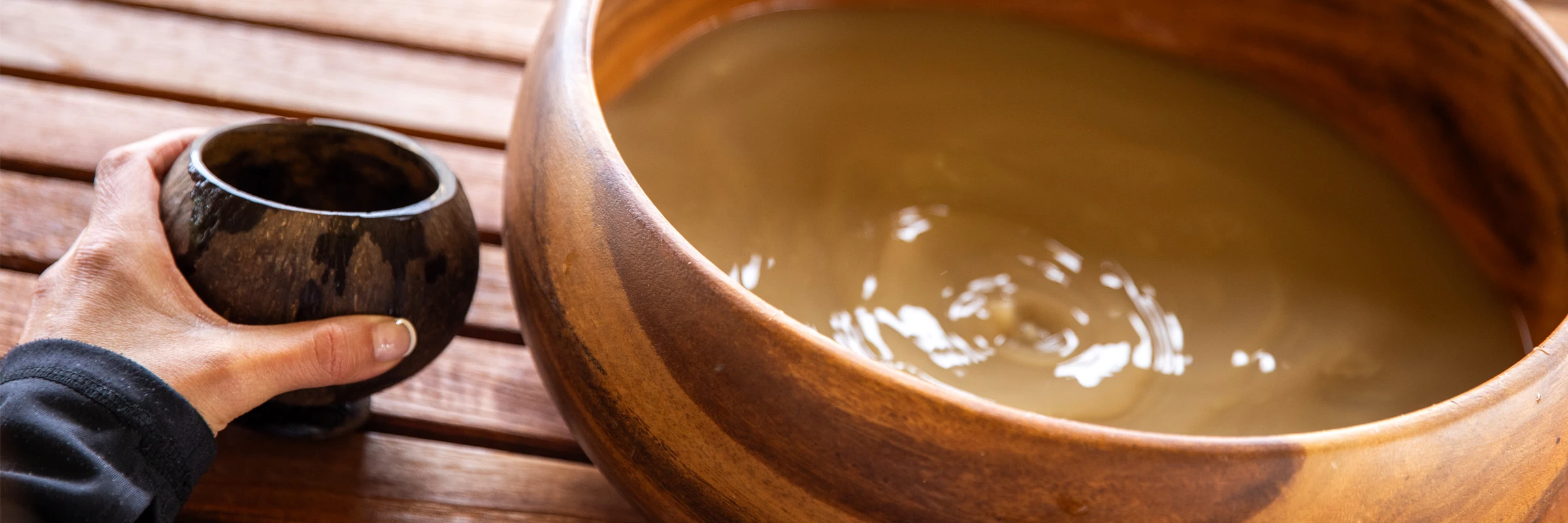 A hand holding a polished coconut-shell ʻapu beside a koa bowl of freshly strained ʻawa.