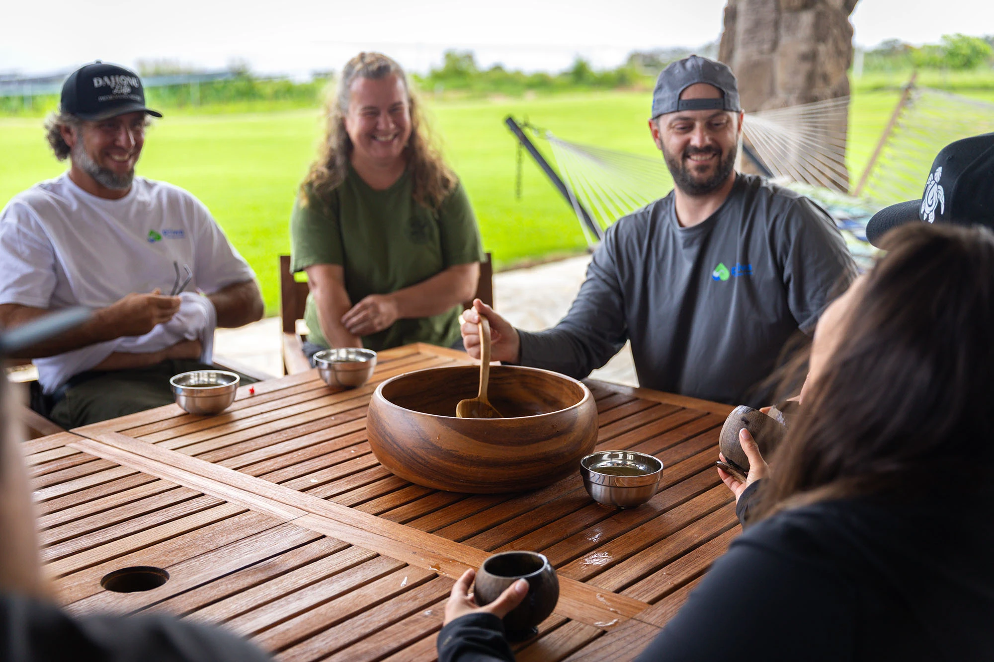 Friends sitting around a wooden table outdoors, sharing ʻawa from a large koa bowl and coconut-shell cups.