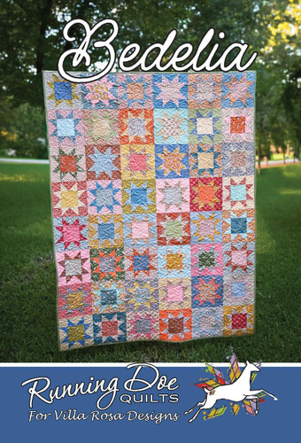 Villa Rosa Designs Bedelia Pattern quilt, colorful patchwork quilt hanging outdoors on a grassy area with trees in the background.