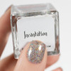 Incantation Nail Polish, close-up of a hand holding a clear square bottle with holographic glitter nail polish and a sparkling, multicolored nail design on the thumb.