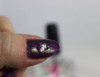 Marvel Liquid Macro Top Coat Nail Polish, close-up of a fingernail with glossy purple nail polish featuring holographic glitter, with nail polish bottle in the background.