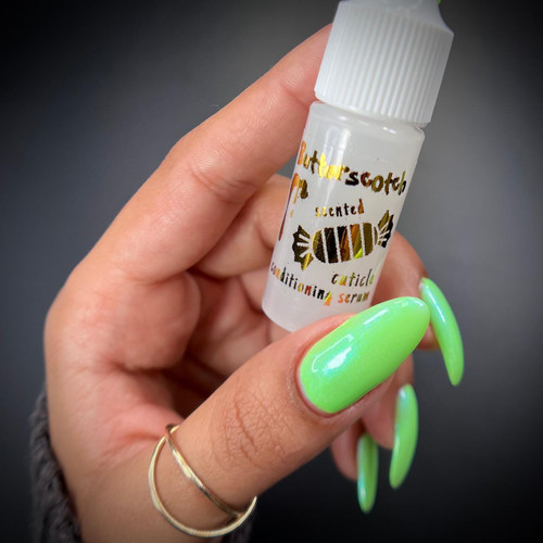 Butterscotch Scented Cuticle Conditioning Serum, small white bottle with colorful label and ribbed cap held by a hand with bright green nails.