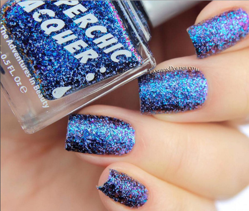 In The Twinkling Of An Eye Nail Polish, close-up of glittery blue nail polish on fingernails with the bottle of the nail polish shown in the background.