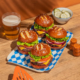 4 hamburgers using King's hawaiian Original Hawaiian Sweet Pretzel Hamburger Buns, with a bowl of pickles, a spatula, and a sparkling beverage on a wood surface