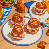 4 ham and cheese sliders on a white plate using King's Hawaiian Original Hawaiian Sweet Pretzel Pre-Sliced Slider Buns with some chips, an orange salt shaken, single King's Hawaiian pretzel bite on wooden table