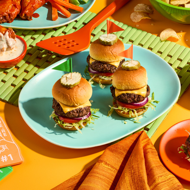 3 Sliders on King's Hawaiian Pre-Sliced Slider Buns on turquoise plate on orange surface surrounded by Buffalo wings potato chips and an orange spatula