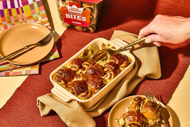 King's Hawaiian Cinnamon Soft Swirl Bites Casserole with tub if bites in the background, a plate of casserole and a fork diving into the casserole on a red table runner