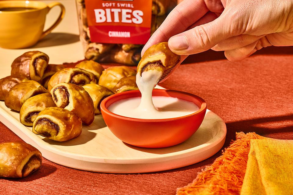 One King's Hawaiian Soft Swirl Cinnamon Bite being dipped into cream cheese frosting in an orange bowl, with a tub of King's Hawaiian Soft Swirl Cinnamon Bites​ and Soft Swirl Cinnamon Bites​ on a plate and a cup of coffee in the background